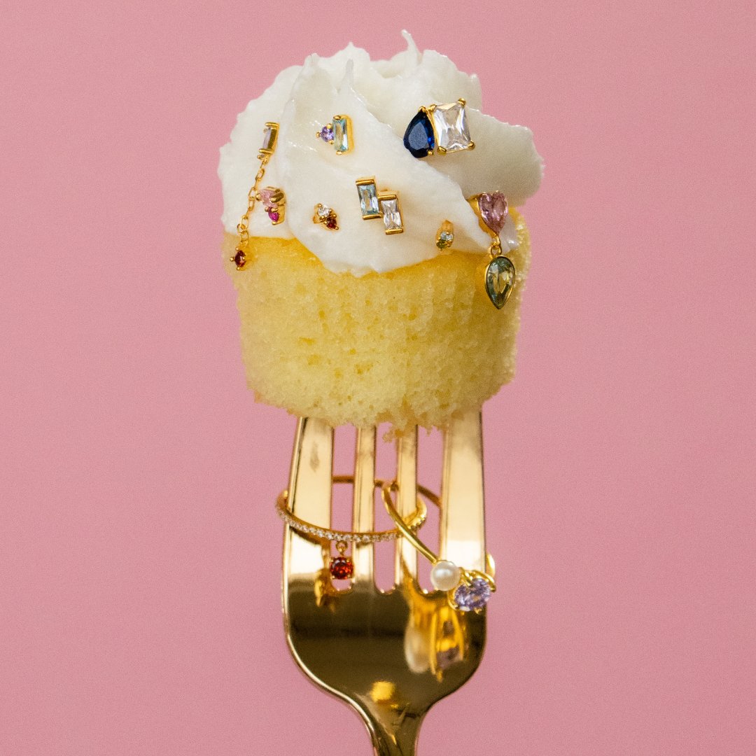 Vanilla cupcake with white frosting and colorful jewelry on a gold fork, against a pink background.