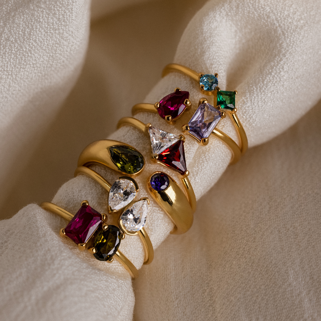 Six gold rings with colorful gemstones displayed on a soft, cream-colored fabric.