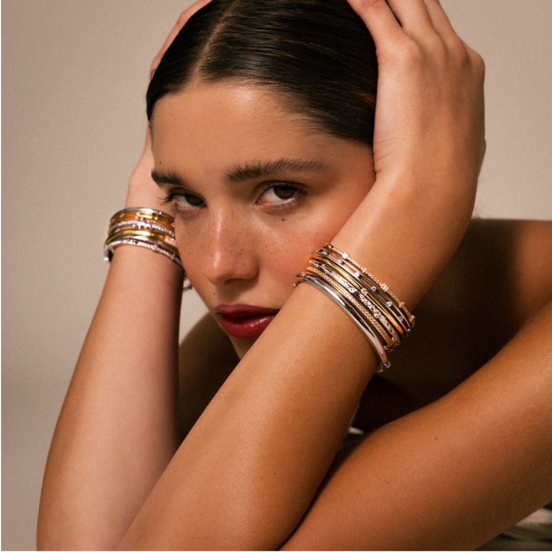 Close up image of a model's wrist showcasing a gold bracelet stack and beaded hand chains over a water background