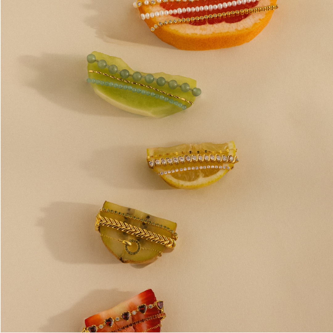 Close up image of a model's wrist showcasing a gold bracelet stack and beaded hand chains over a water background