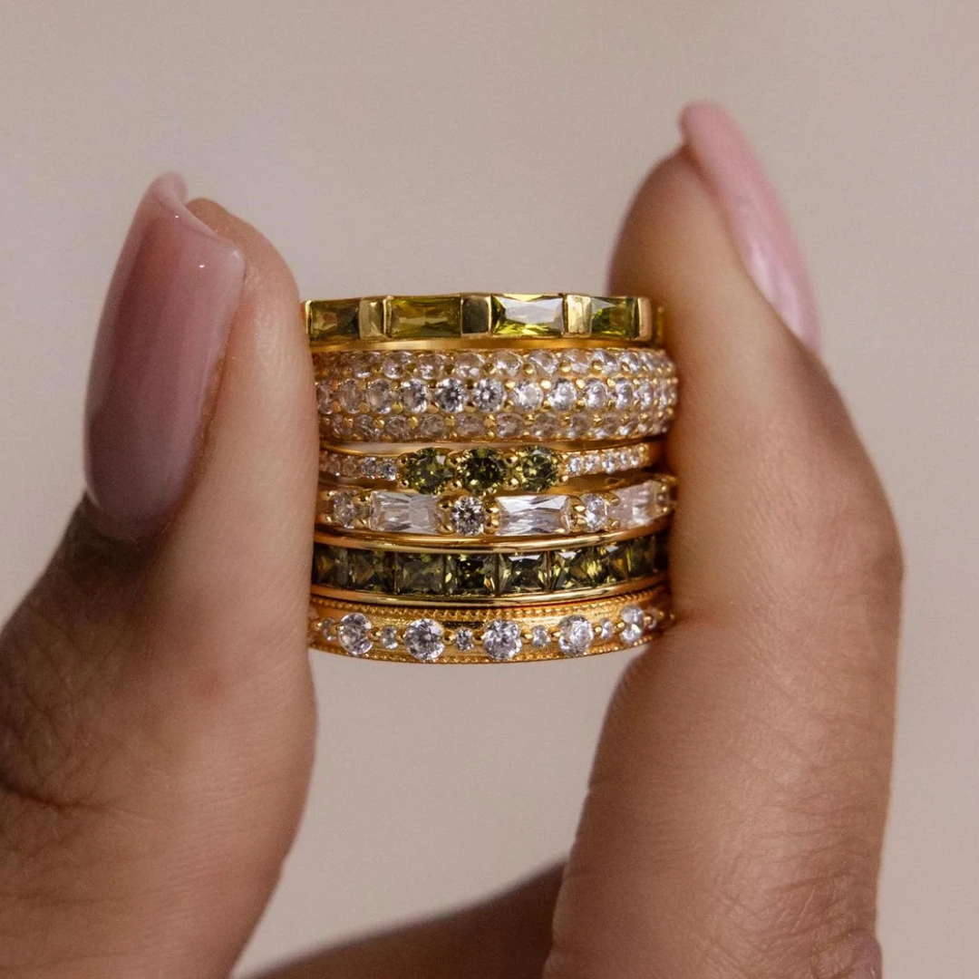 A hand with manicured nails holds a stack of gold and diamond rings against a neutral background.