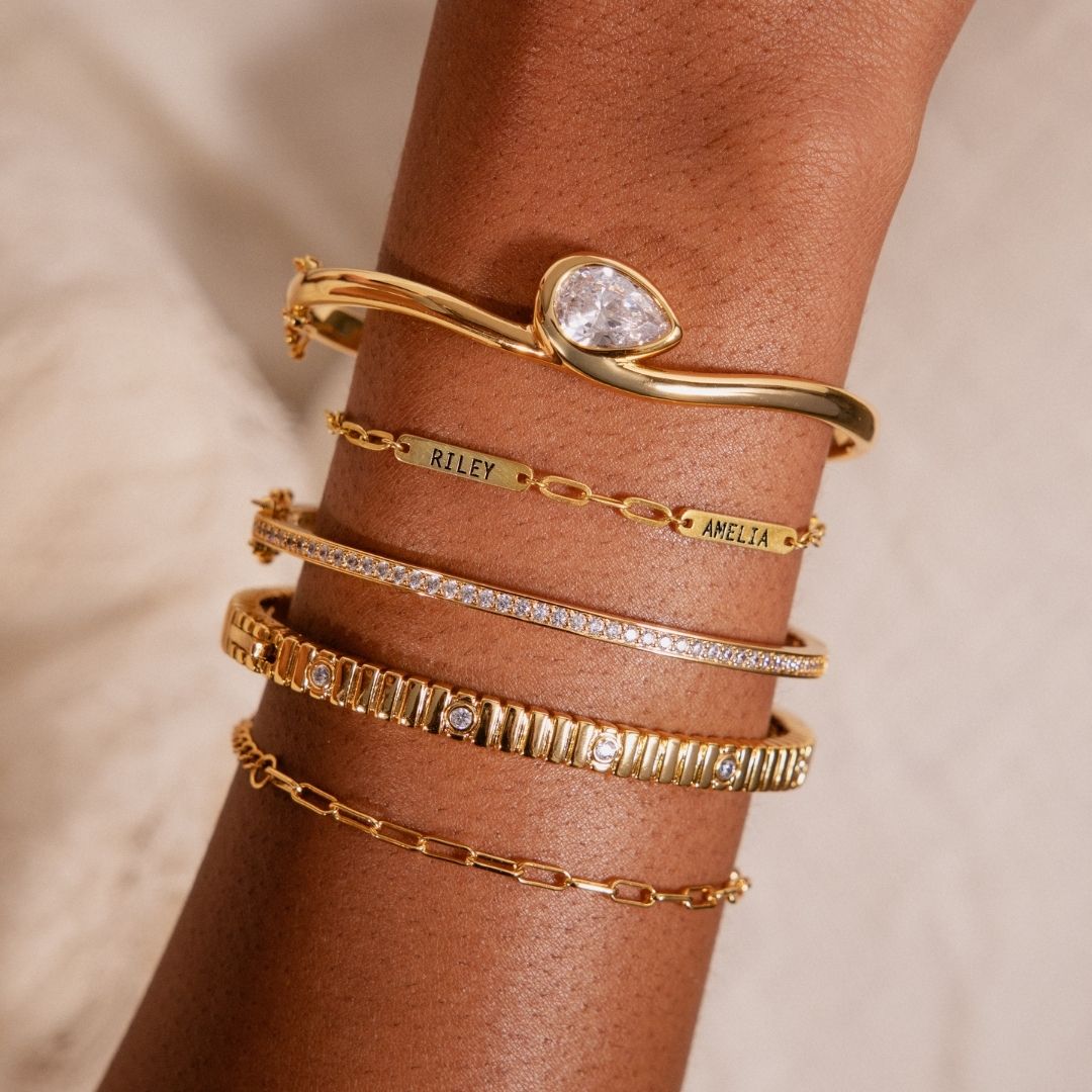 Close up image of a model's wrist showcasing a gold bracelet stack and beaded hand chains over a water background