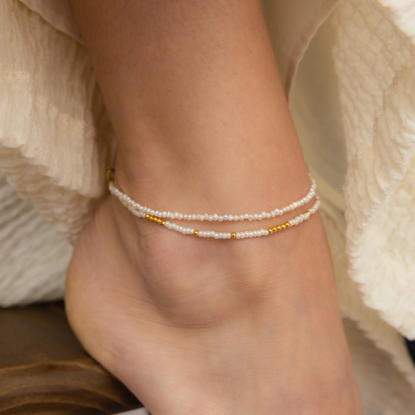 A close-up of an ankle wearing two delicate beaded anklets with gold accents.