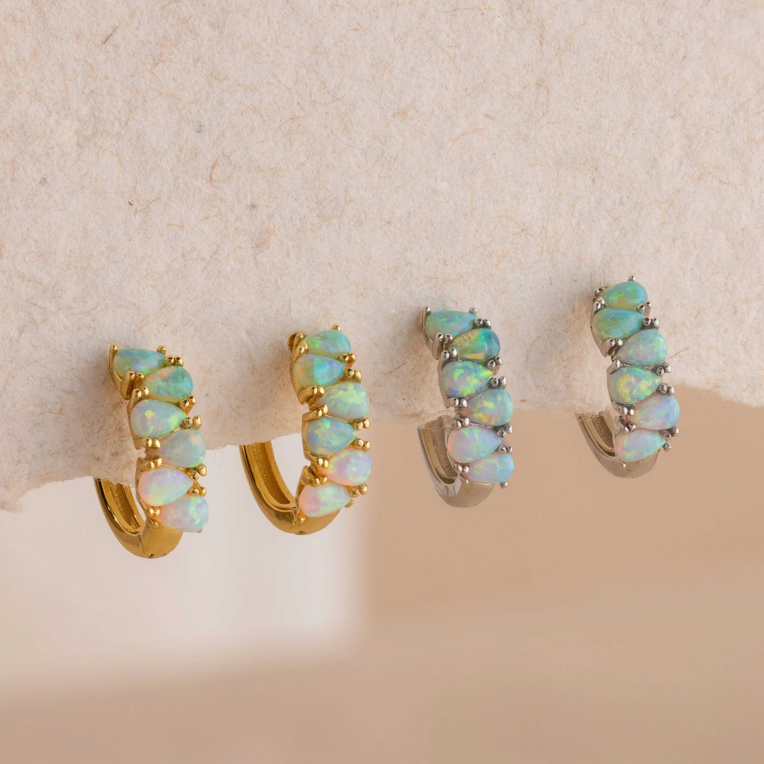 Four opal hoop earrings, two gold and two silver, displayed on a neutral textured surface.