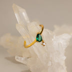 The Duo Birthstone Drop Ring, featuring a green teardrop gem and a small diamond charm in gold, is displayed on clear quartz crystals.