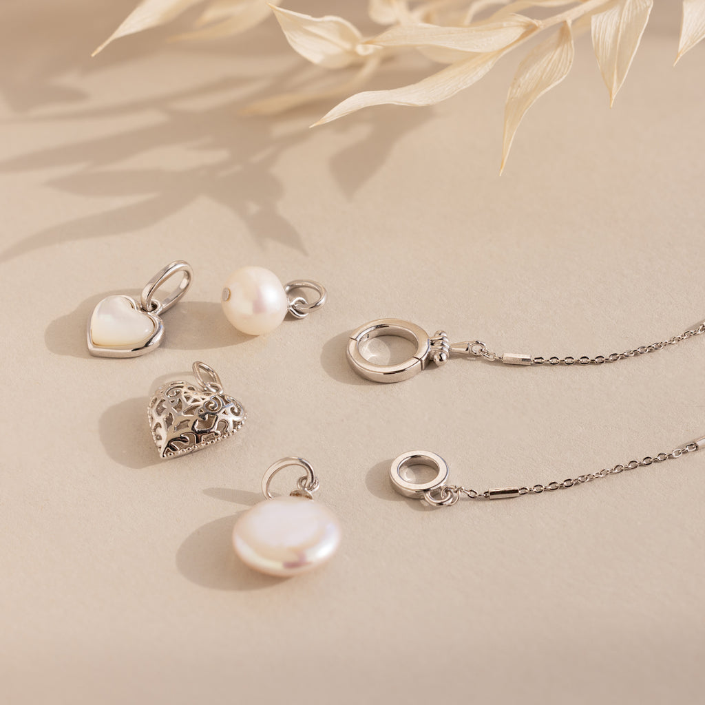 The Hinged Circle Charm Bracelet in Sterling Silver is styled with silver necklaces and pendants featuring pearl and heart designs, all displayed on a beige background with dried leaves for a minimal jewelry aesthetic.
