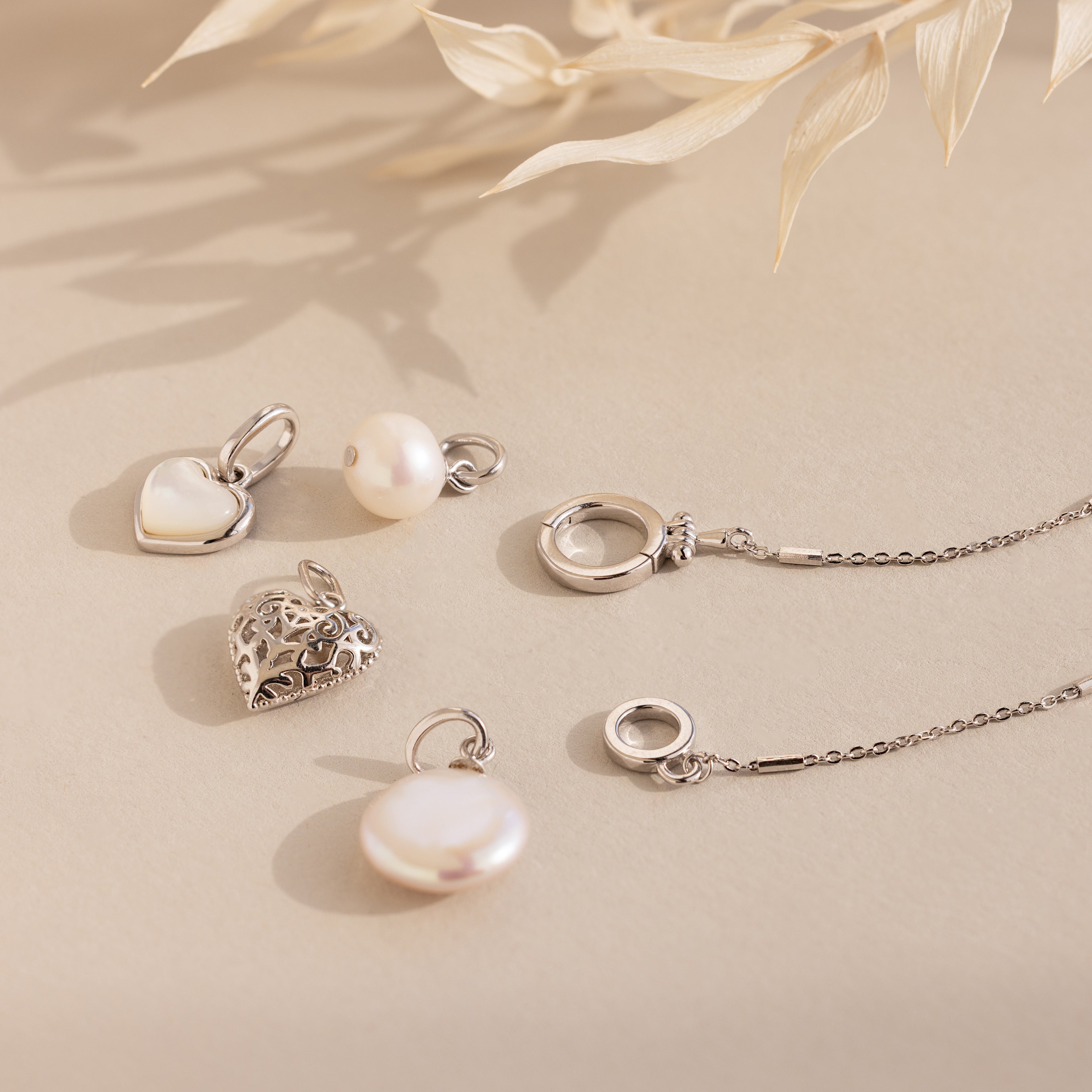 The Hinged Circle Charm Bracelet in Sterling Silver is styled with silver necklaces and pendants featuring pearl and heart designs, all displayed on a beige background with dried leaves for a minimal jewelry aesthetic.