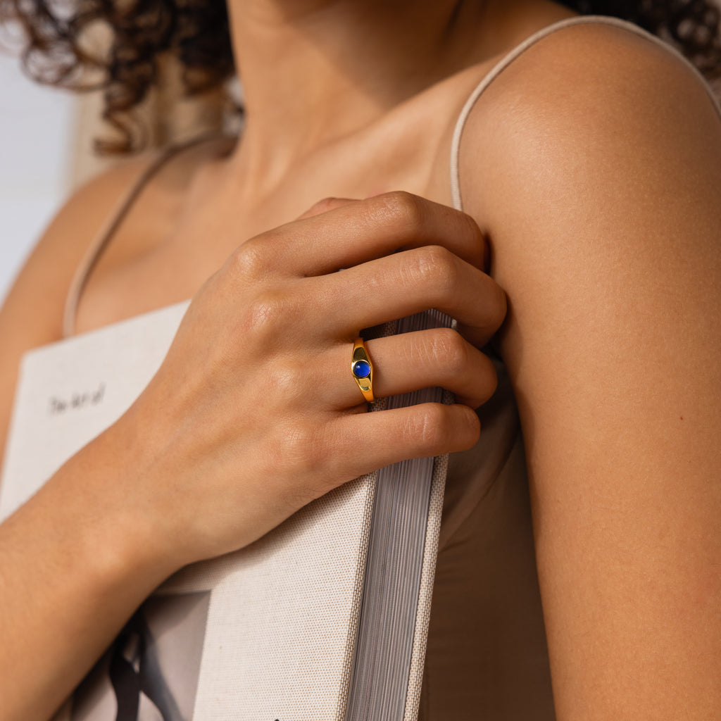 A person in a beige top holds a book, wearing nostalgic jewelry—a gold Signet Mood Rings Set with a blue stone—on their finger.