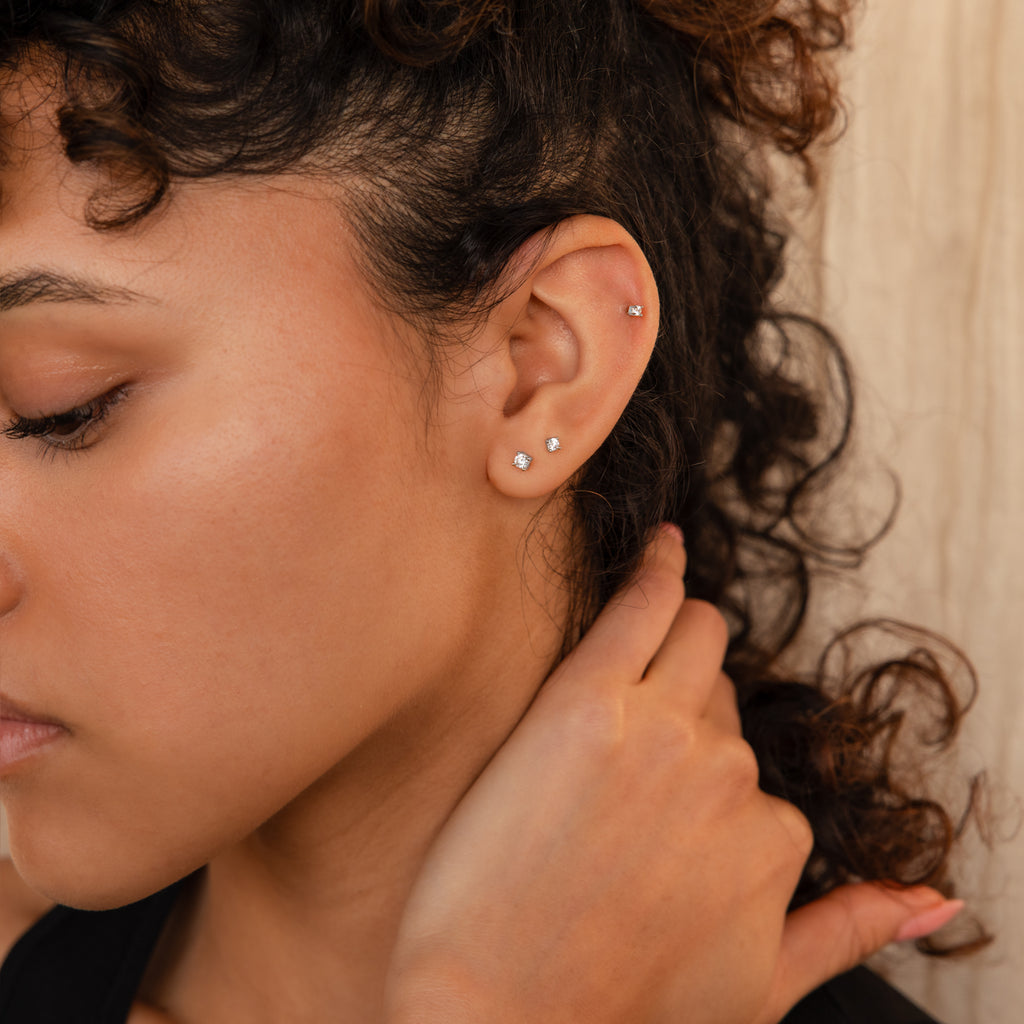 A woman with curly hair displays three dainty earrings, including the Diamond Flatback Studs Set in Sterling Silver, in her ear as she gently touches her neck.