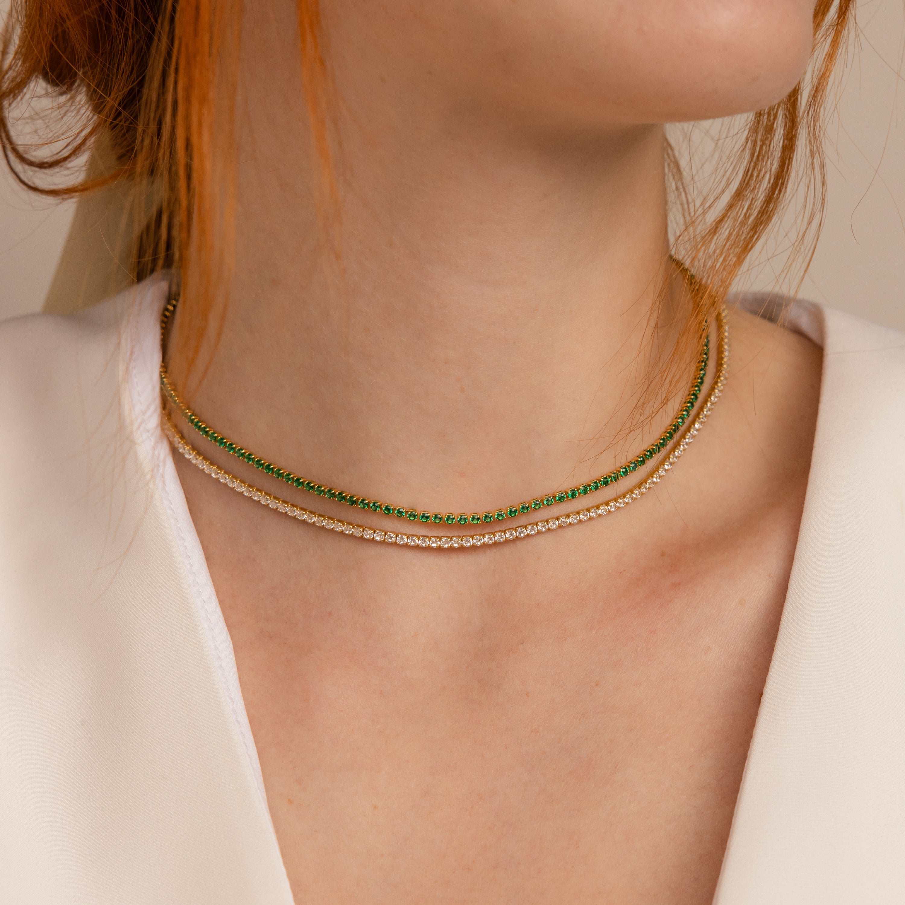 Layered necklace style featuring a gold tennis choker with green stones paired with a gold tennis choker with clear stones, worn with a white v-neck top.