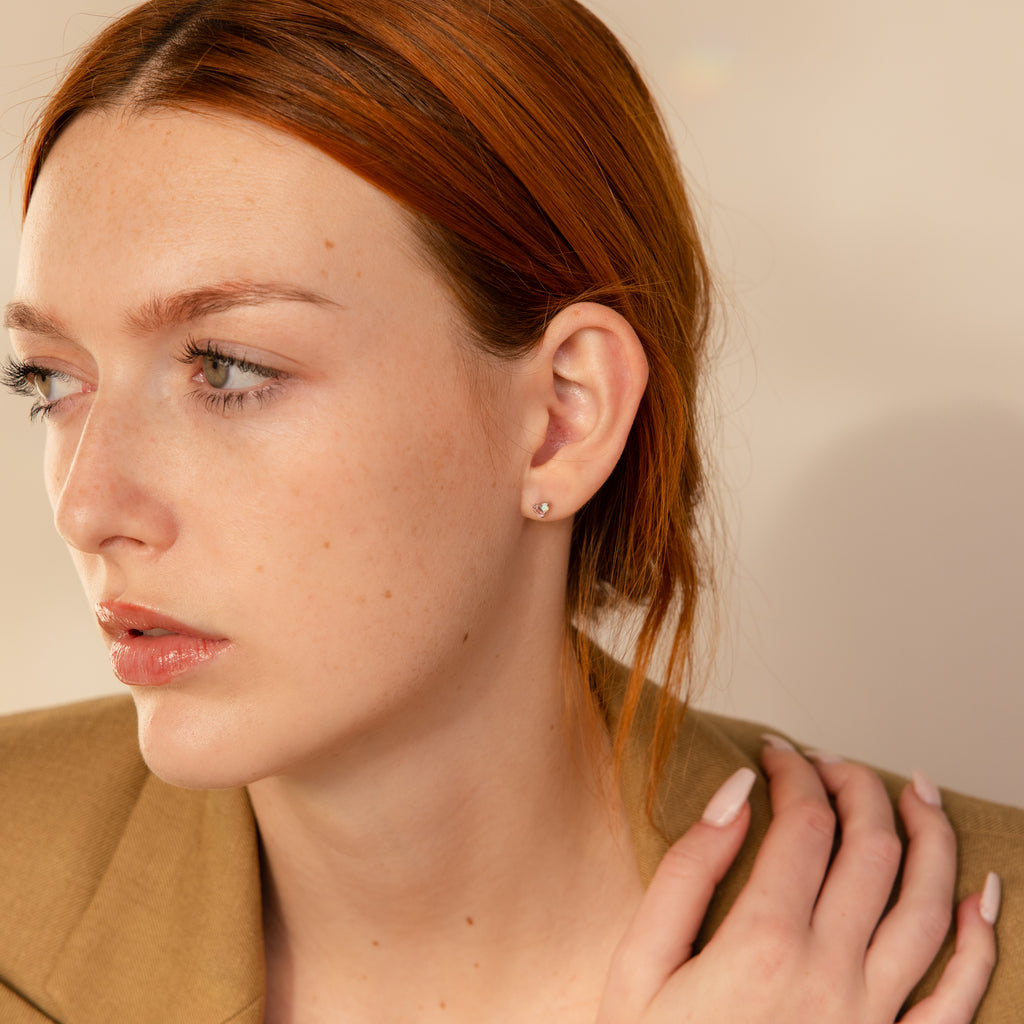 A red-haired, fair-skinned woman in a tan blazer gazes thoughtfully, wearing Pink Baguette Opal Studs in Silver—an ideal bridesmaid gift.