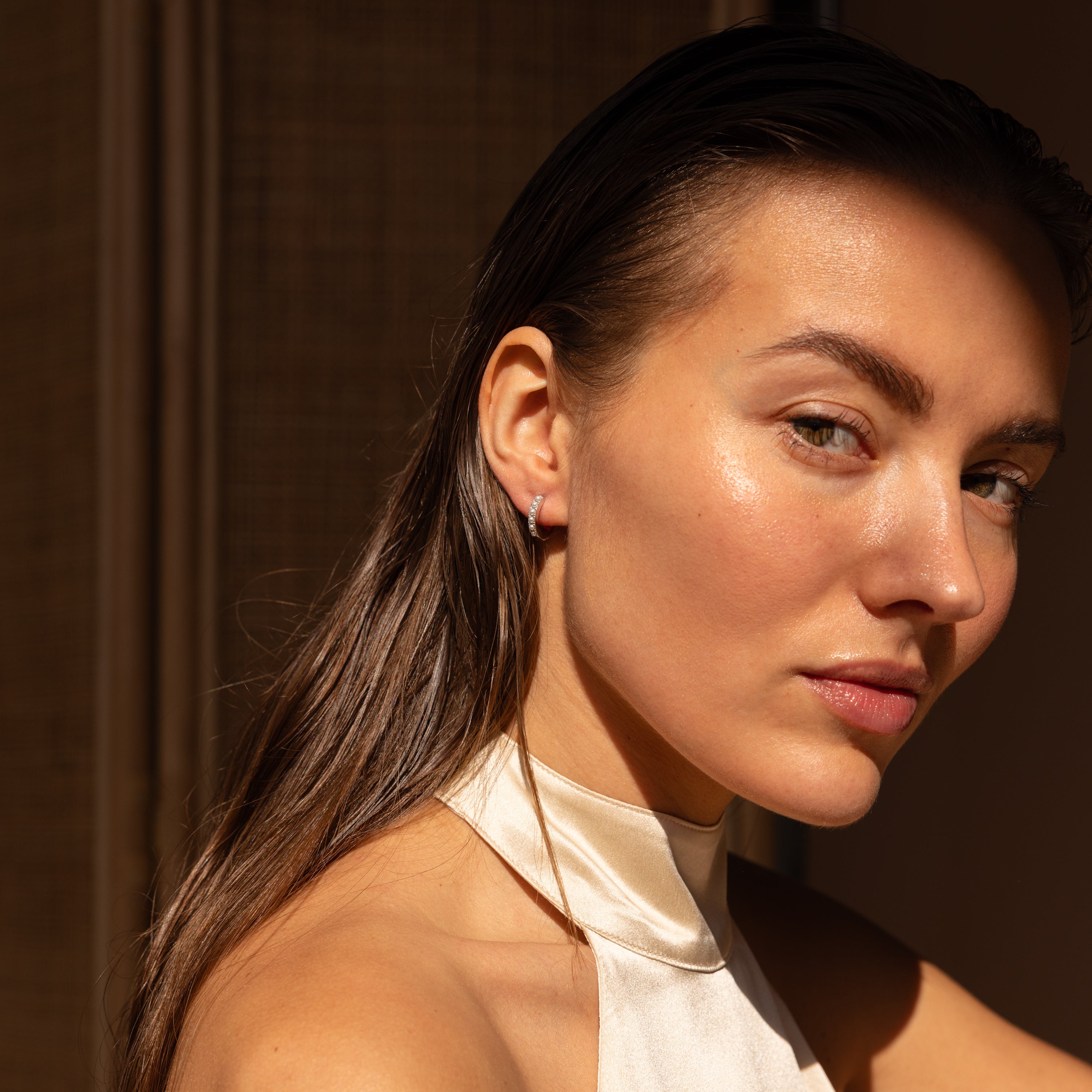 Woman with wet hair in a satin halter top, gazing confidently in warm natural light, her look elevated by elegant Perle Huggies in Sterling Silver—the perfect wedding accessory.