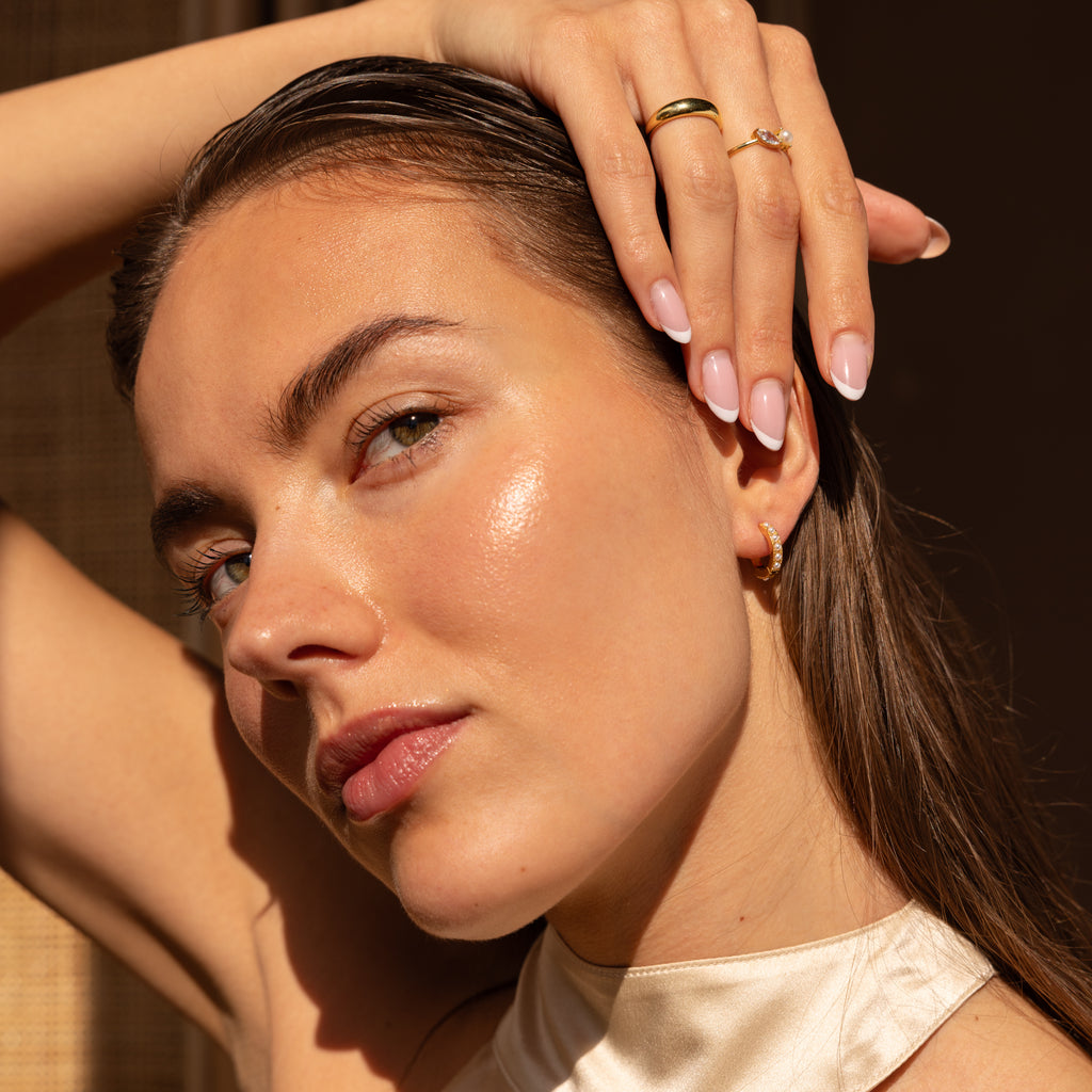 A woman wearing a pair of gold huggie earrings adorned with small pearl-like beads. Her hair is slicked back, and she has manicured nails and stacked gold rings, creating a polished and elegant look.