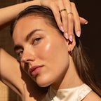 A woman wearing a pair of gold huggie earrings adorned with small pearl-like beads. Her hair is slicked back, and she has manicured nails and stacked gold rings, creating a polished and elegant look.