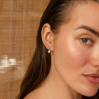 A woman with wet hair faces the camera against a woven beige background, wearing Dainty Knot Pearl Earrings in Sterling Silver, radiating timeless sophistication.