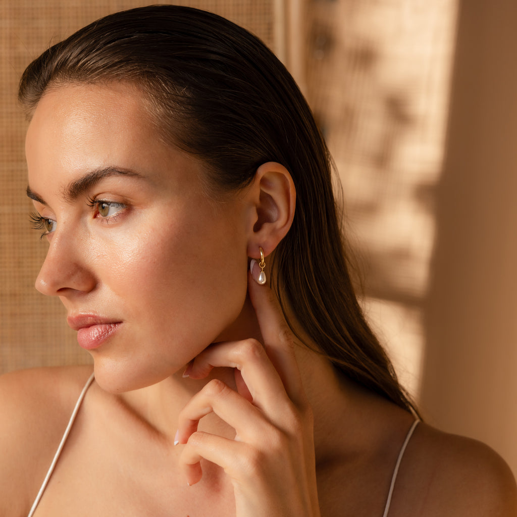 Woman with long brown hair touches her Dainty Pearl Drop Huggies in 18K Gold, looking to the side in soft natural light.