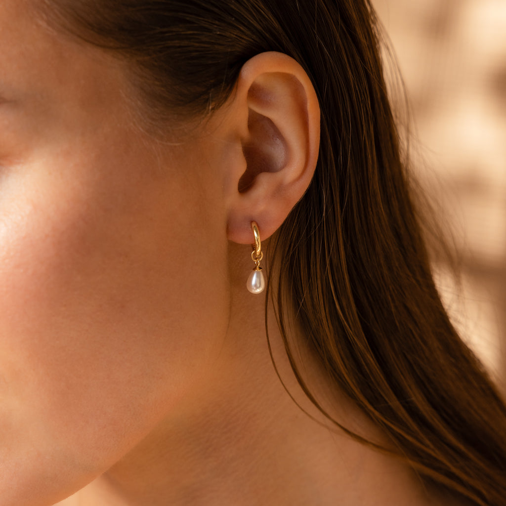Close-up of a woman wearing Dainty Pearl Drop Huggies—gold hoops with hanging pearls—set against a soft, neutral background.