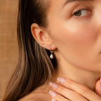 A woman wearing Dainty Pearl Drop Huggies in Sterling Silver gently touches her chin with manicured fingers.