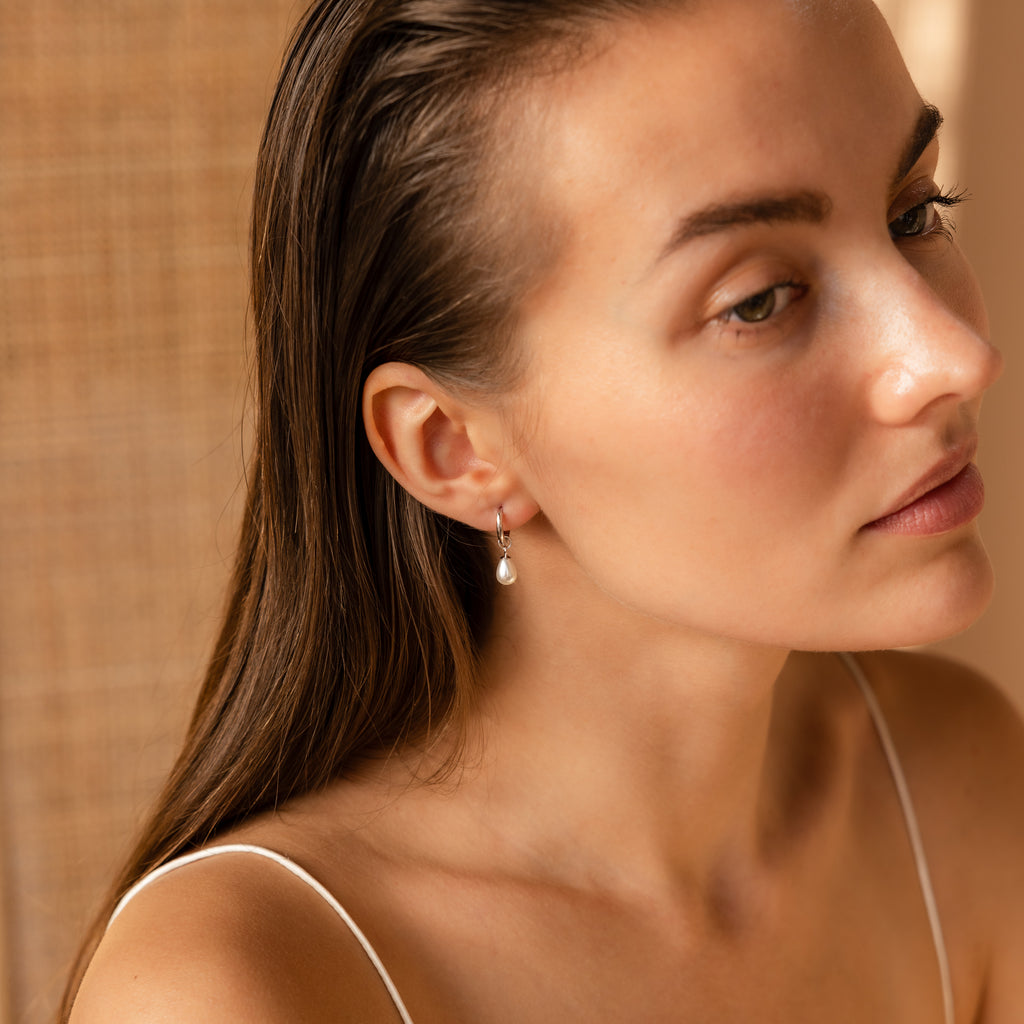 A woman with wet hair, wearing a white top and Dainty Pearl Drop Huggies in Sterling Silver, gazes to the side in soft lighting.