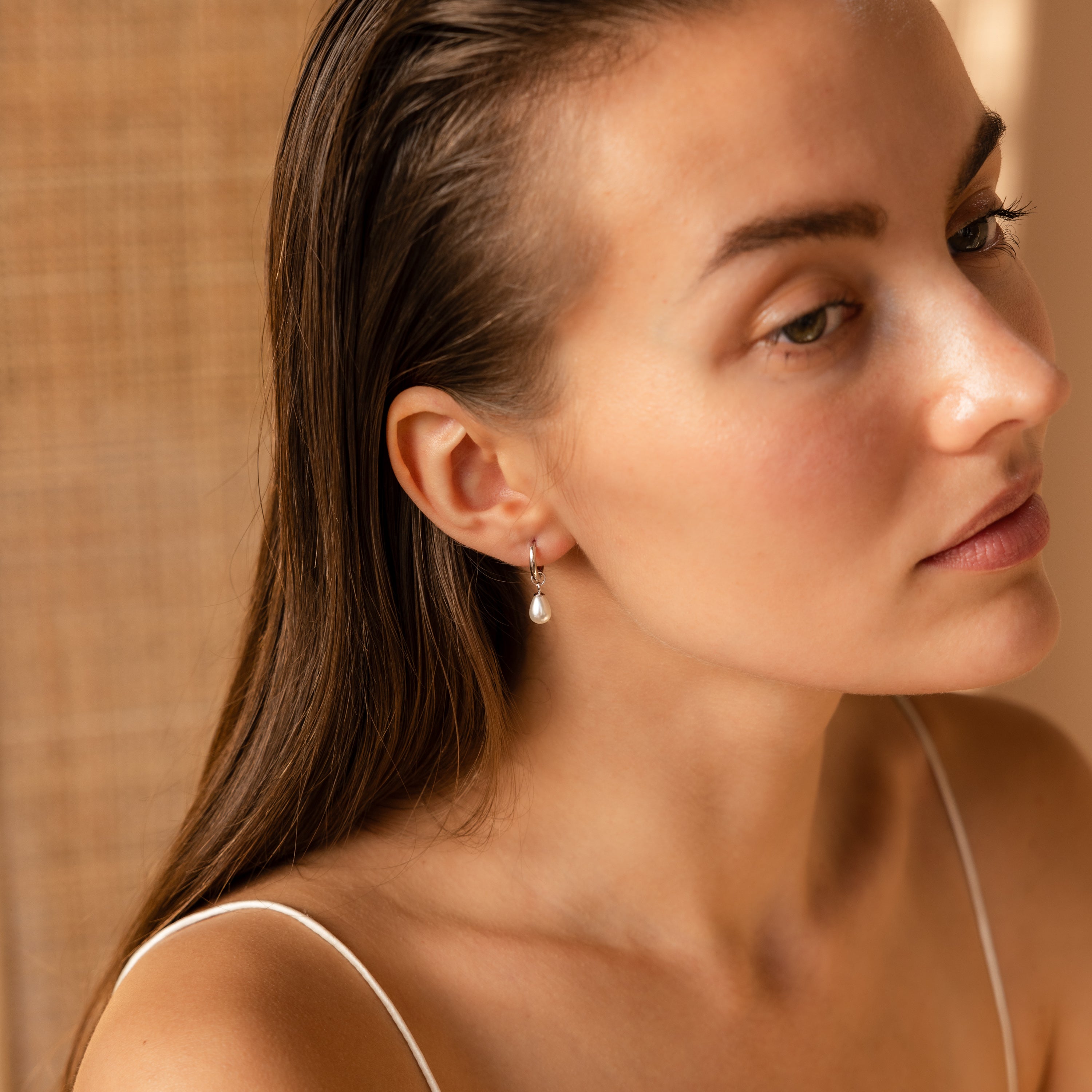 A woman with wet hair, wearing a white top and Dainty Pearl Drop Huggies in Sterling Silver, gazes to the side in soft lighting.