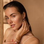 A woman with wet hair touches her earring and looks at the camera, showcasing the Dainty Pearl Drop Huggies and a stylish pearl bracelet.