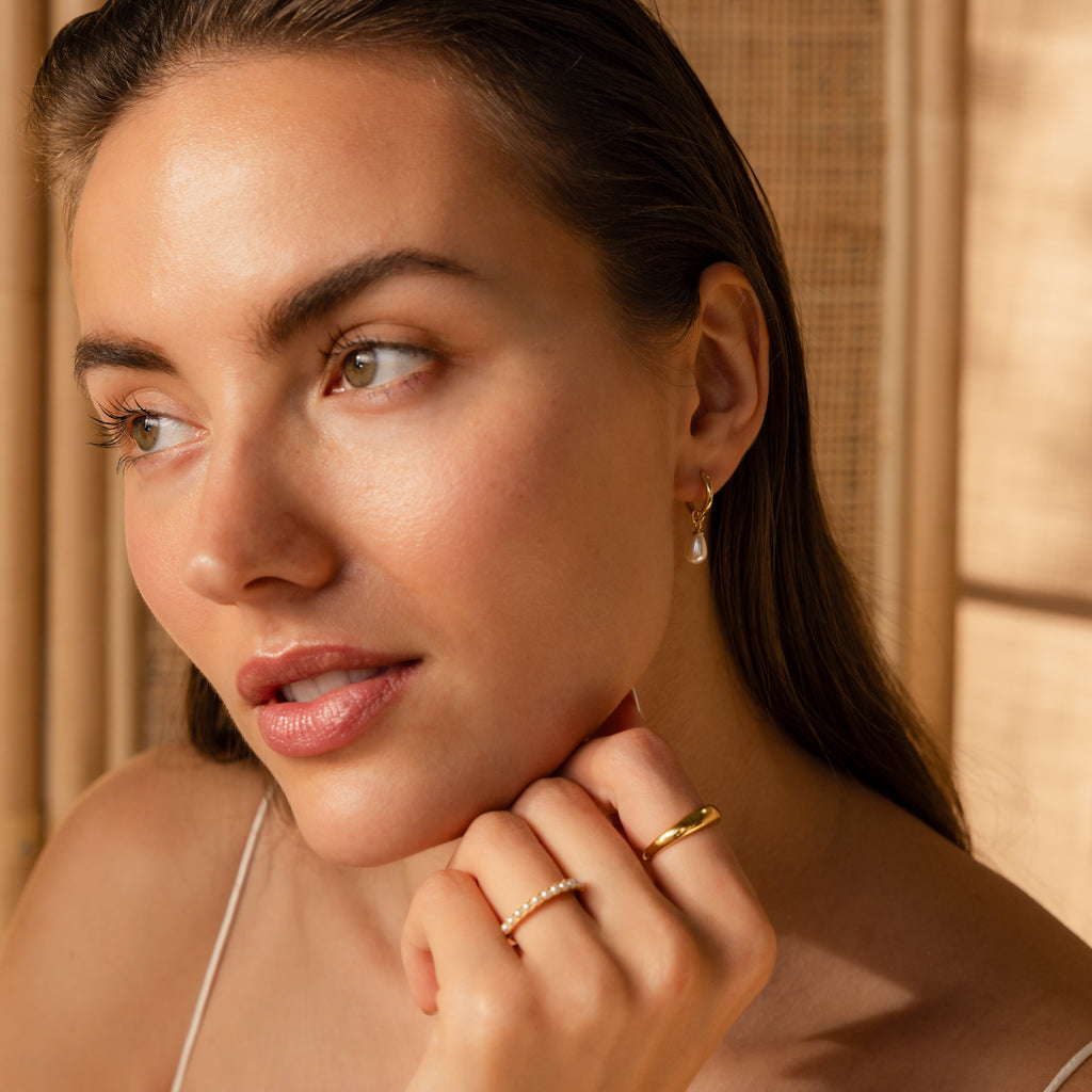Woman with light makeup wears gold rings and Dainty Pearl Drop Huggies in 18K Gold, gazing thoughtfully to the side.