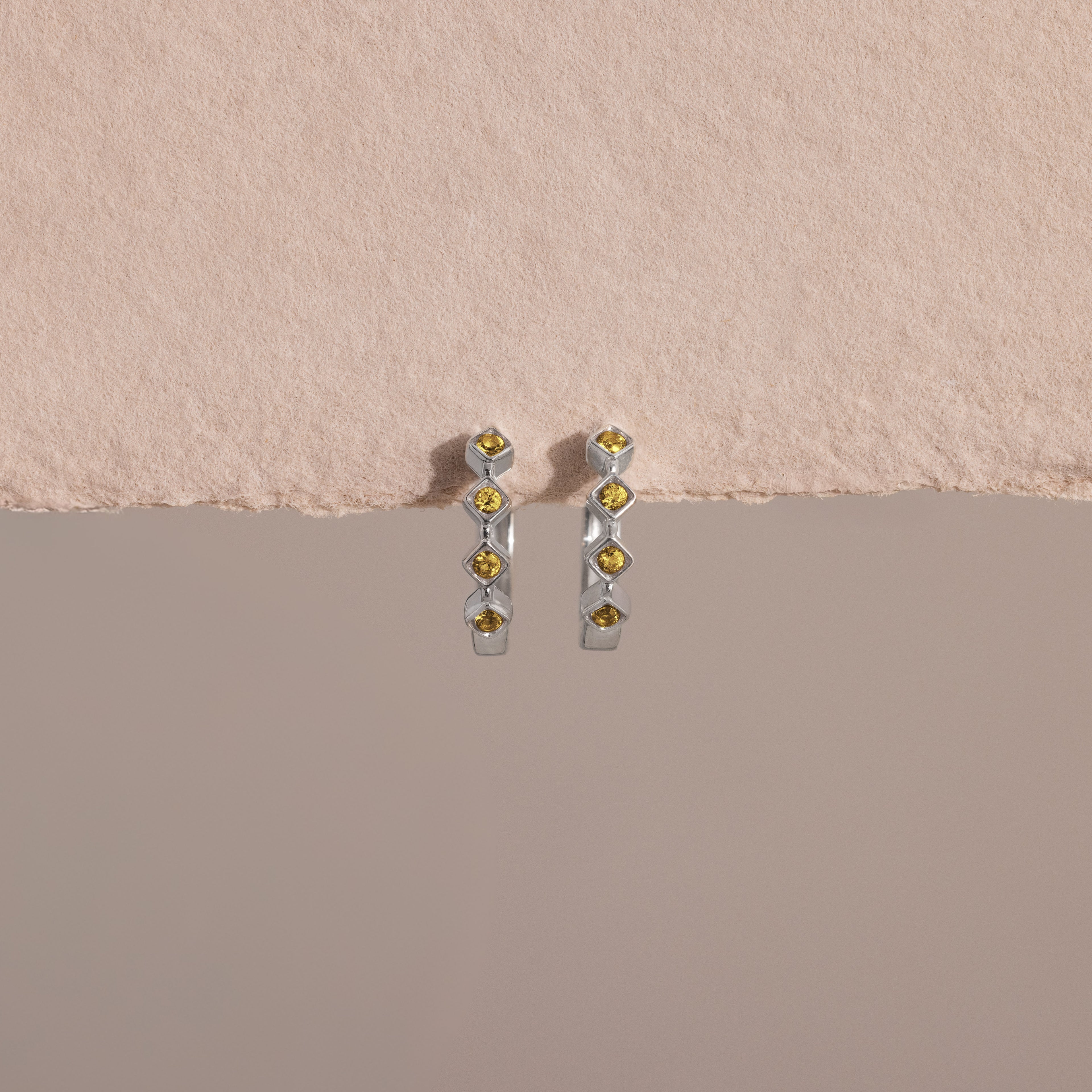 Geometric Topaz Huggies—silver earrings featuring five yellow topaz stones each, inspired by geometric birthstone jewelry, shown on a textured light pink background.
