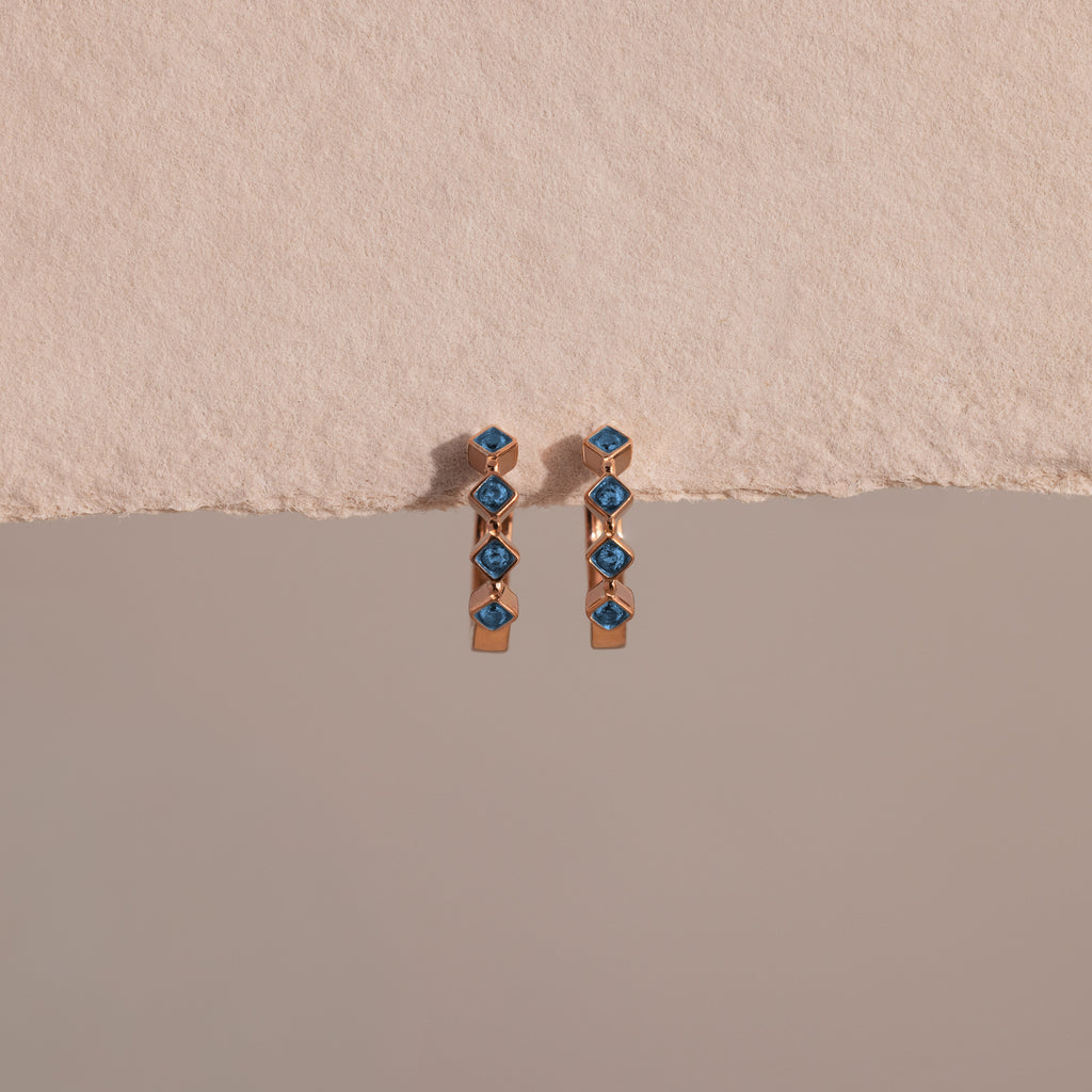 Two Geometric Blue Zircon Huggies, crafted in gold with blue zircon gemstones and delicate diamond accents, are displayed against a textured beige and smooth gray background.