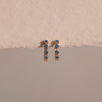 Two Geometric Blue Zircon Huggies, crafted in gold with blue zircon gemstones and delicate diamond accents, are displayed against a textured beige and smooth gray background.