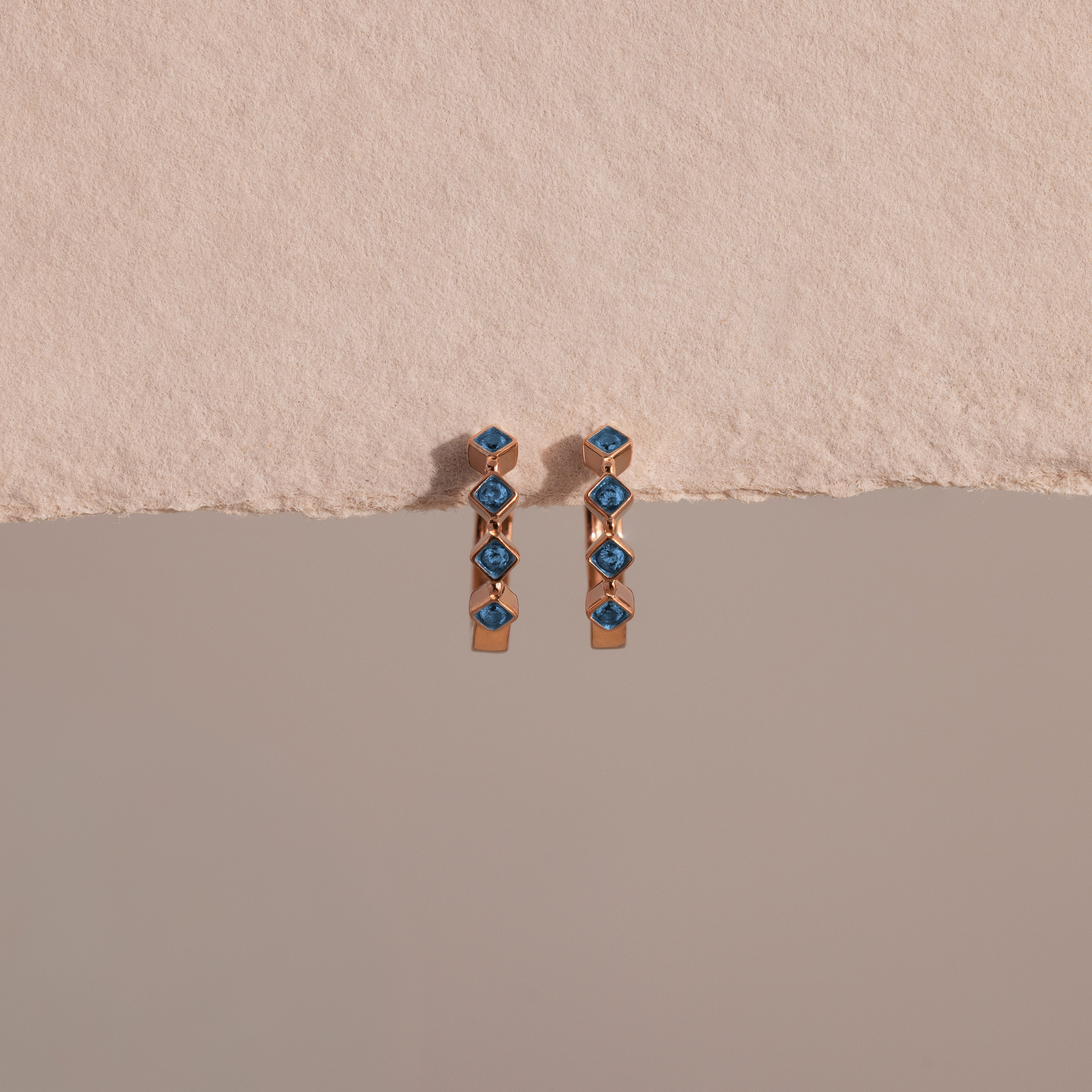 Two Geometric Blue Zircon Huggies, crafted in gold with blue zircon gemstones and delicate diamond accents, are displayed against a textured beige and smooth gray background.
