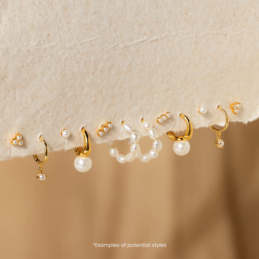 Mystery Jewelry gold and pearl earrings in assorted styles, each with whimsical charm, elegantly showcased against a cream fabric background.
