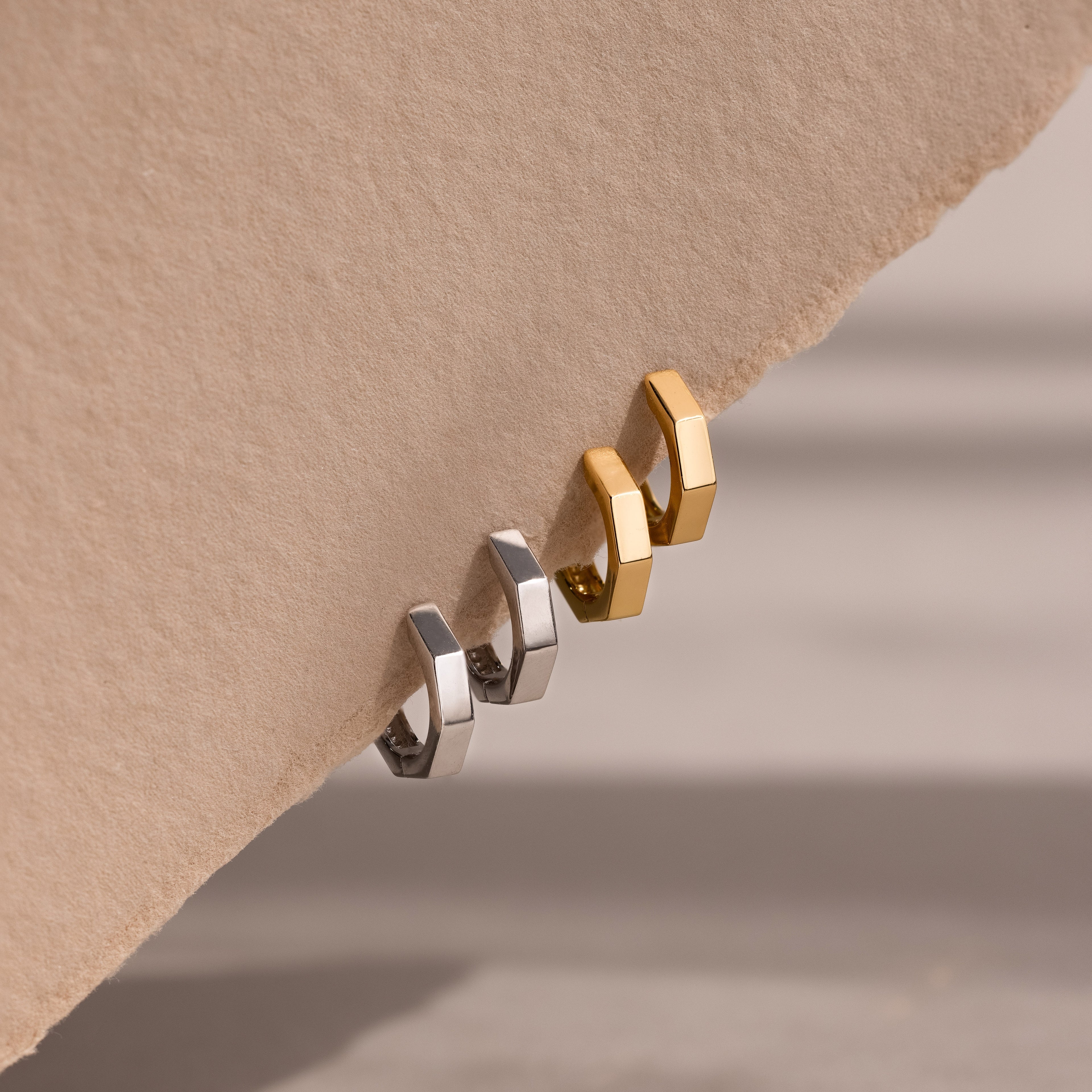 Two pairs of gold and silver geometric hoop earrings on a beige background
