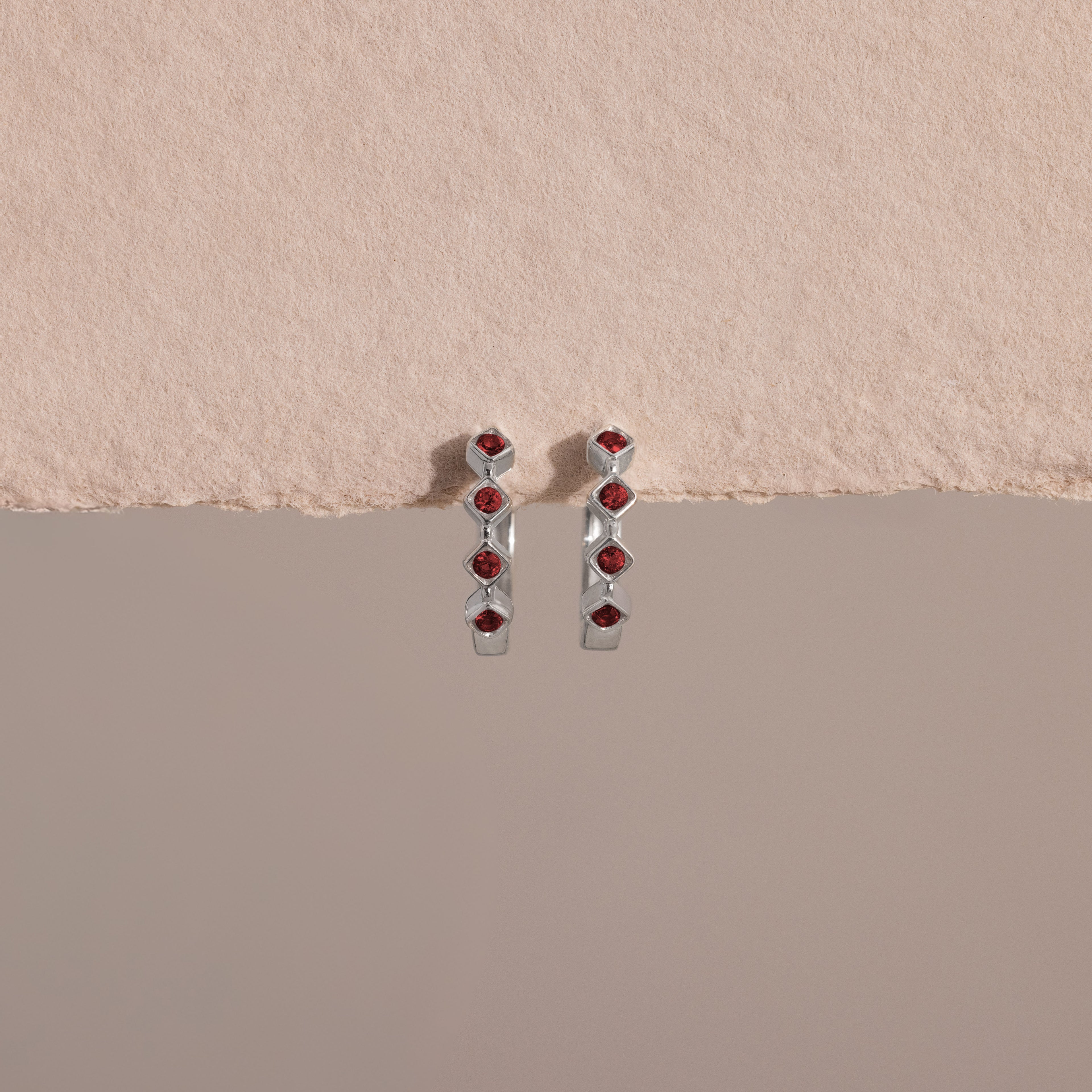 Geometric Garnet Huggies feature silver earrings with small red gemstones arranged vertically, adding a modern touch to your birthstone jewelry collection, showcased on a textured beige surface.