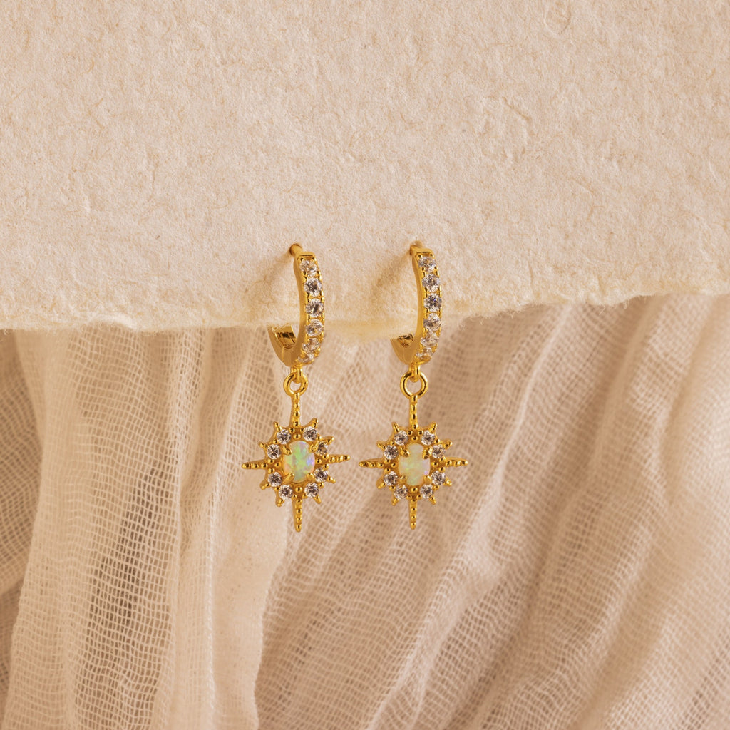 The Opal Star Necklace & Earrings Set features gold hoops with star-shaped opal-like pendants and small clear stones, perfect for pairing with your favorite layered looks. Shown elegantly displayed on textured fabric.