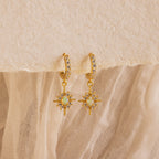 The Opal Star Necklace & Earrings Set features gold hoops with star-shaped opal-like pendants and small clear stones, perfect for pairing with your favorite layered looks. Shown elegantly displayed on textured fabric.