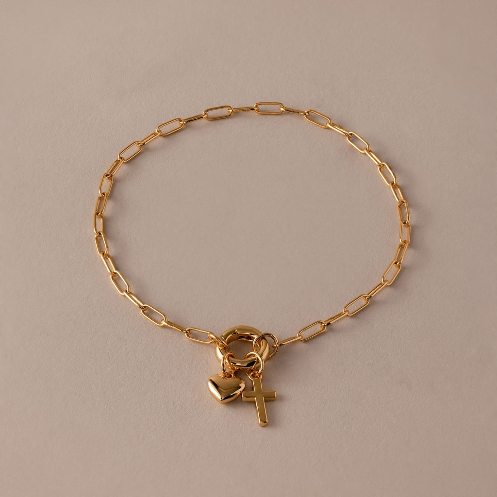 The Cross & Heart Charm Bracelet in 18K Gold features a minimalist cross and heart on a gold paperclip chain, displayed on a beige background.