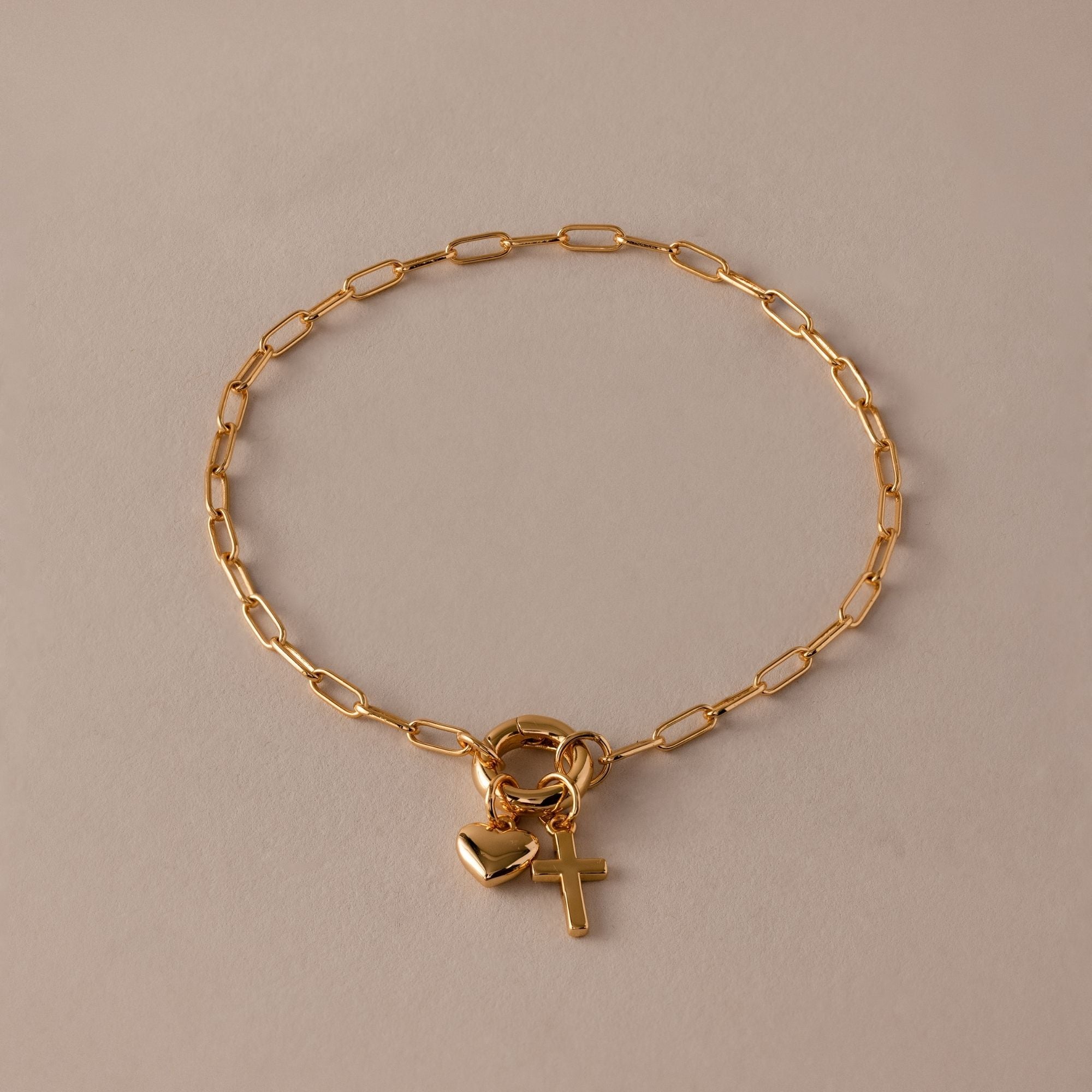 The Cross & Heart Charm Bracelet in 18K Gold features a minimalist cross and heart on a gold paperclip chain, displayed on a beige background.