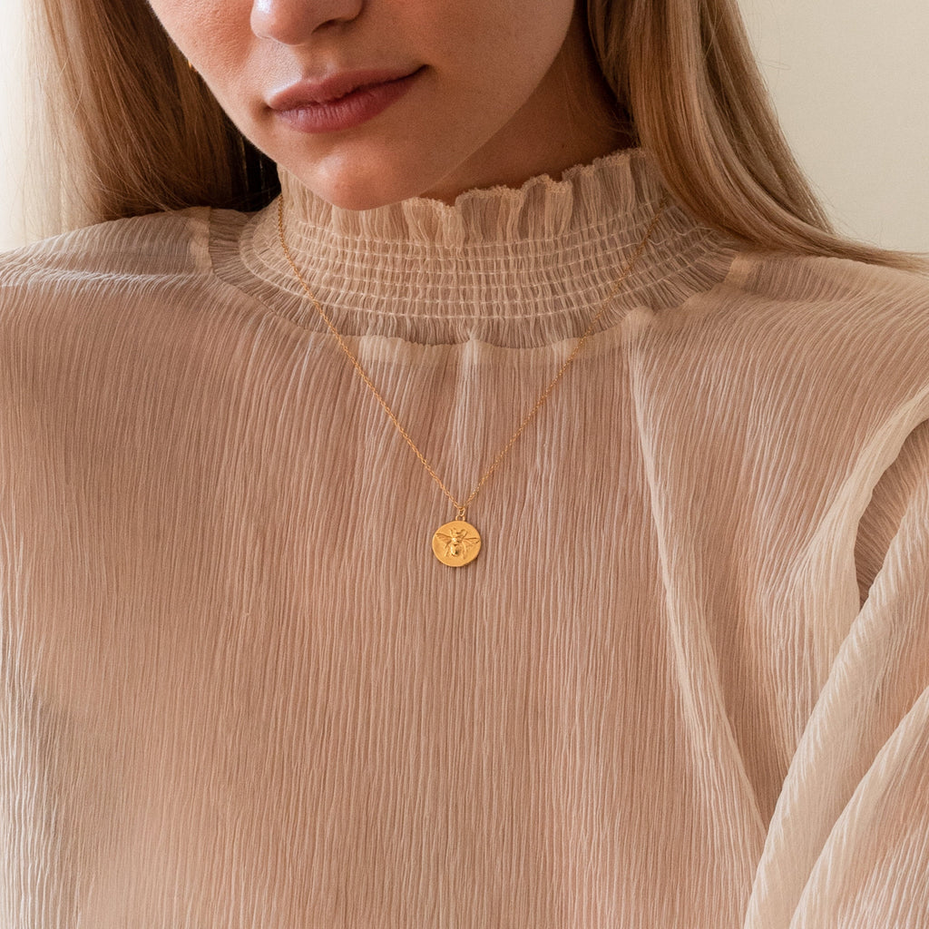 A woman wears a ruffled high-neck beige blouse paired with the Bee Coin Necklace, featuring an engraved gold pendant—perfect for layering with other styles.