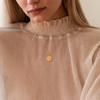 A woman wears a ruffled high-neck beige blouse paired with the Bee Coin Necklace, featuring an engraved gold pendant—perfect for layering with other styles.