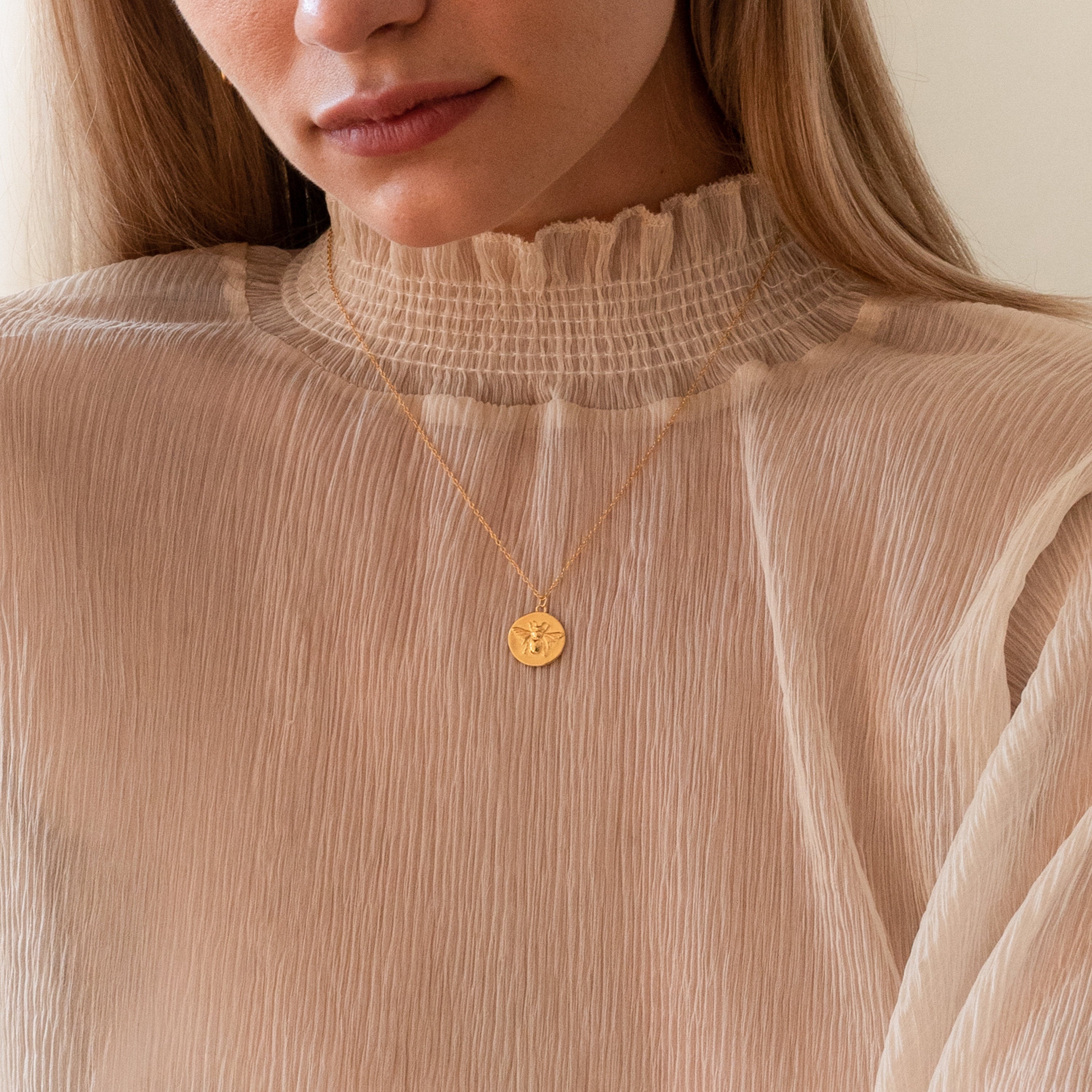 A woman wears a ruffled high-neck beige blouse paired with the Bee Coin Necklace, featuring an engraved gold pendant—perfect for layering with other styles.
