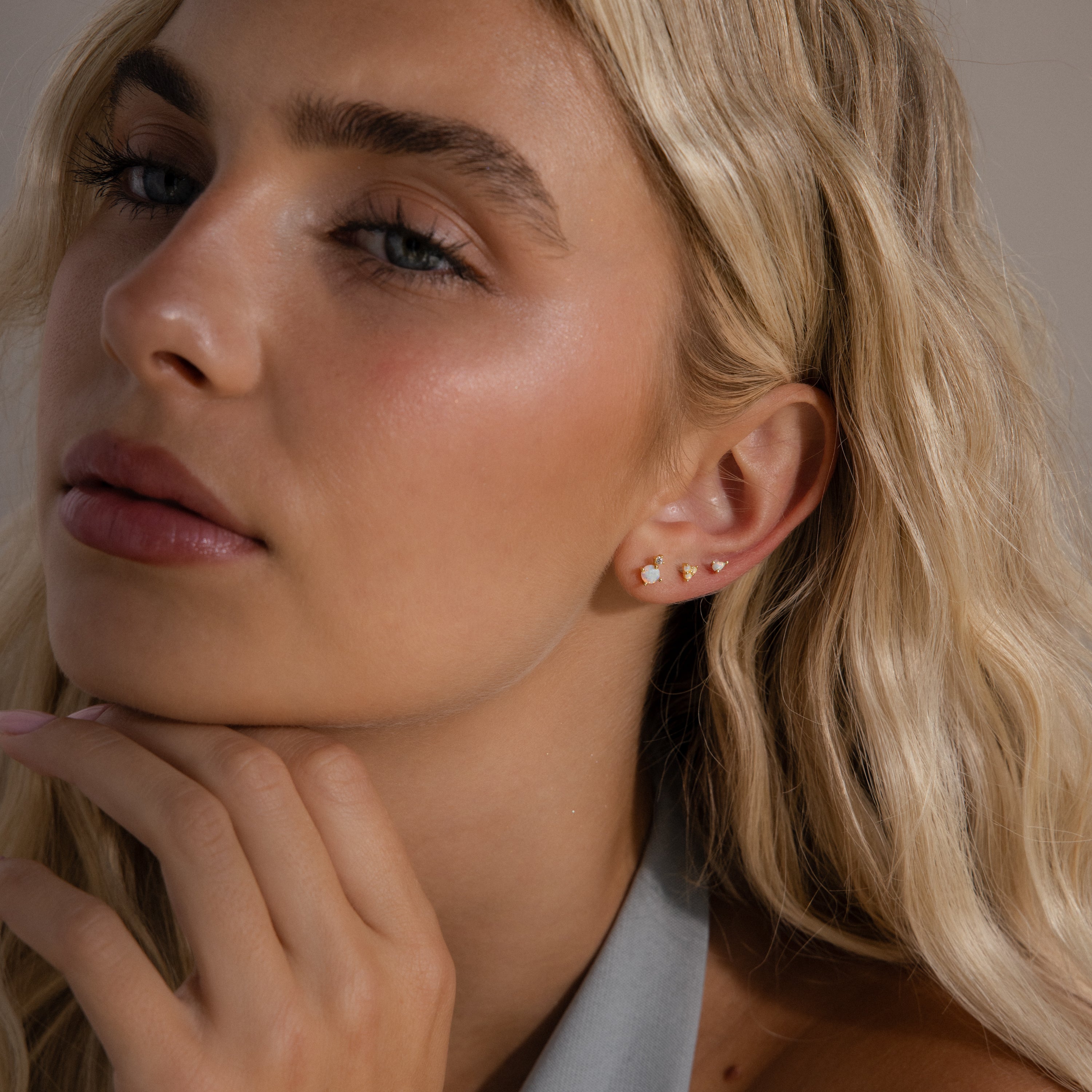 A woman with long blonde hair gently touches her chin, looking thoughtful while wearing two gold earrings and an Opal  Diamond Flatback Stud in 18K Gold.