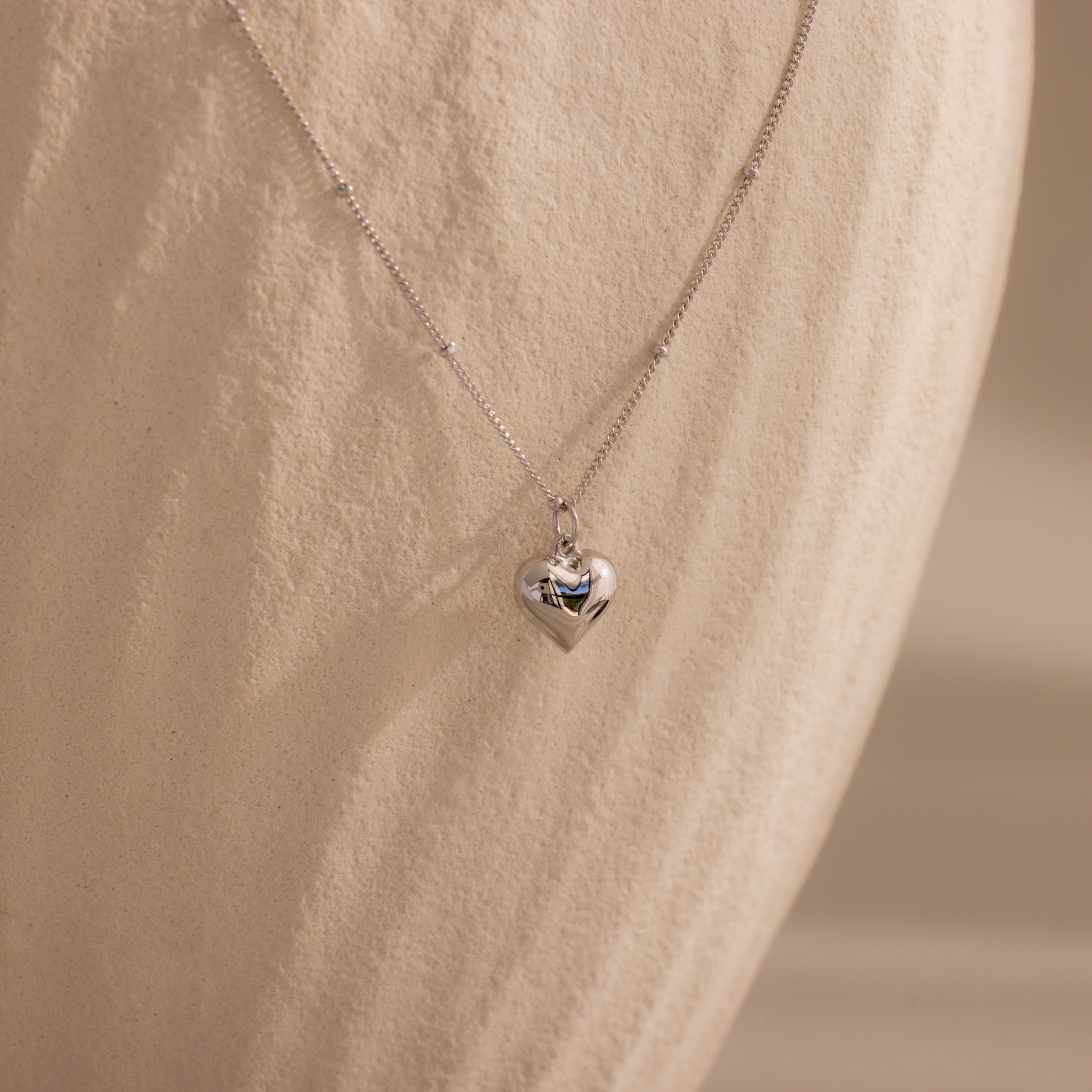 Silver Puff Heart Pendant Necklace displayed against a textured, ivory-toned background.