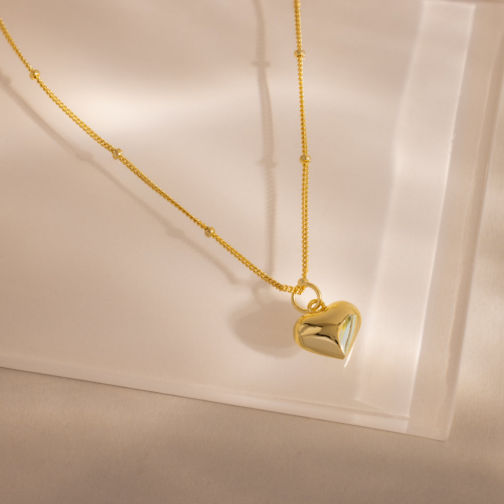 Gold necklace with a puffed heart charm and delicate beaded chain, styled on a soft neutral surface.
