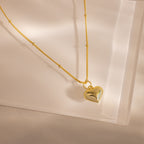 Gold necklace with a puffed heart charm and delicate beaded chain, styled on a soft neutral surface.