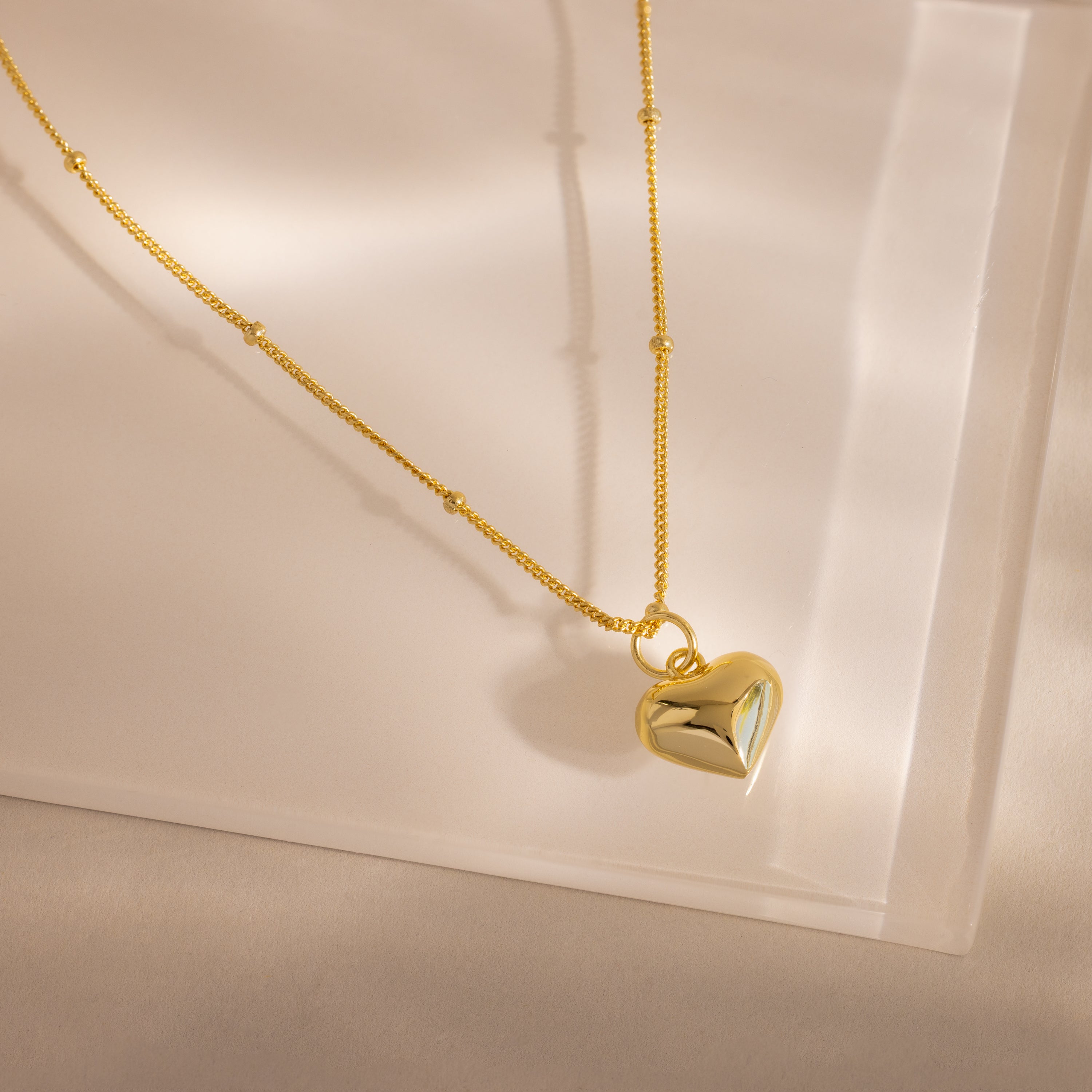 Gold necklace with a puffed heart charm and delicate beaded chain, styled on a soft neutral surface.