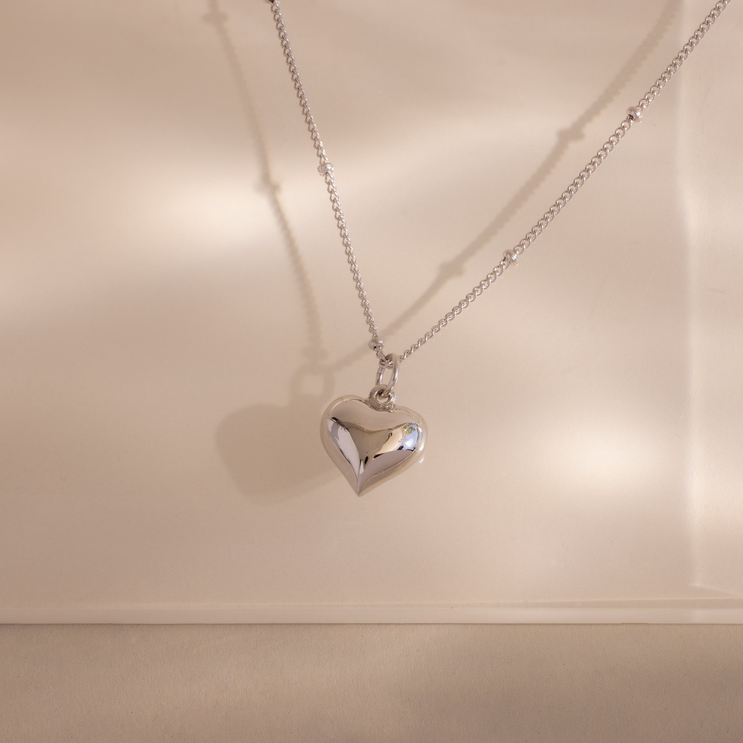 The Puff Heart Charm Necklace in Silver features a silver heart pendant on a delicate satellite chain, creating a subtle shadow against a neutral background.