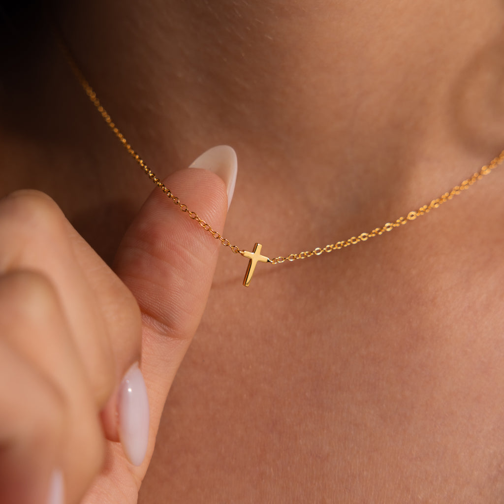 Model wearing a dainty gold gross charm necklace, holding the chain up with her finger.