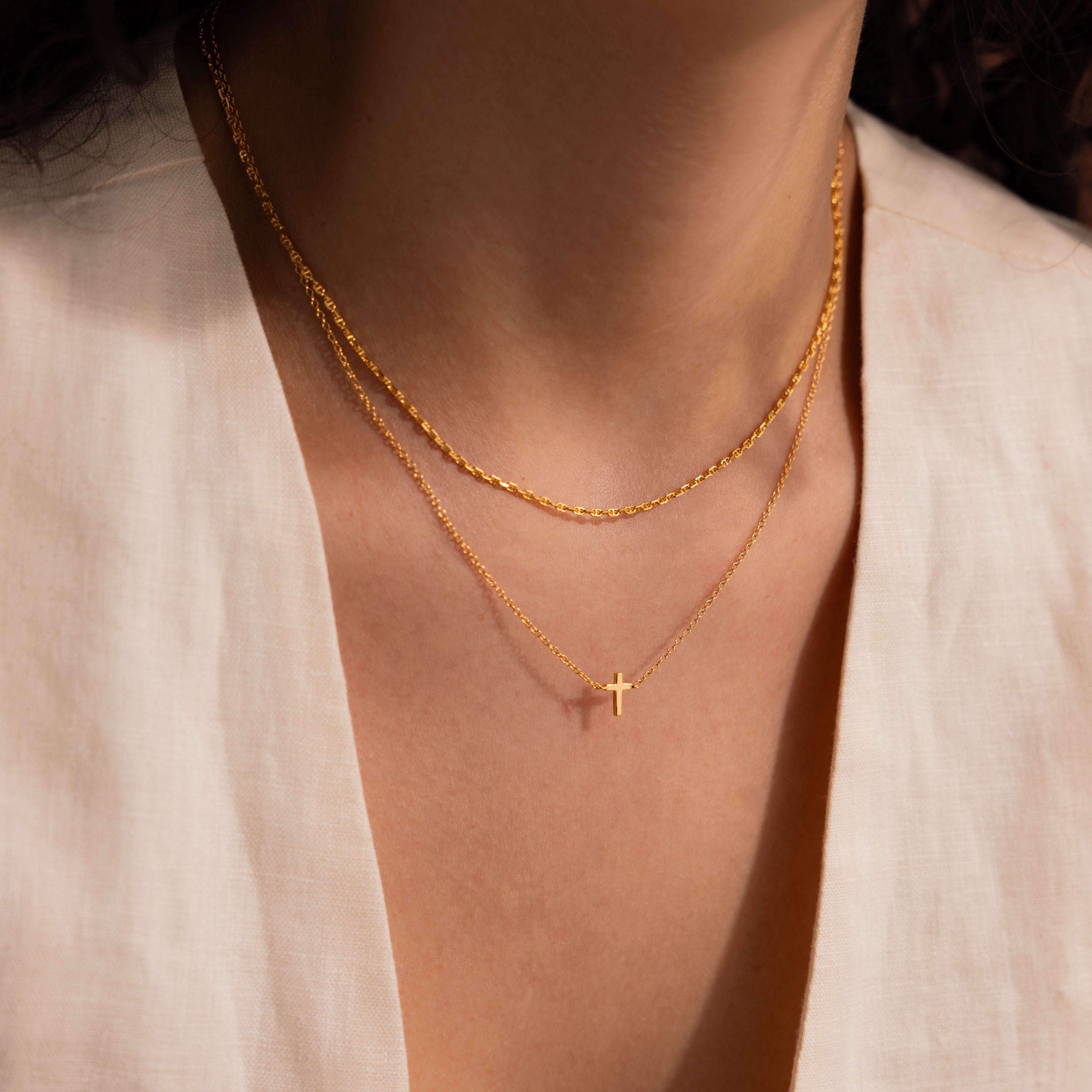 Model wearing a dainty gold cross charm necklace, layered with a gold mariner chain necklace.