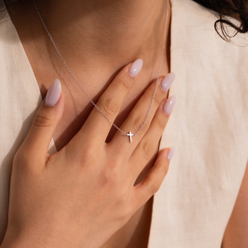 A woman with manicured nails holds the Dainty Cross Necklace pendant near her collarbone.