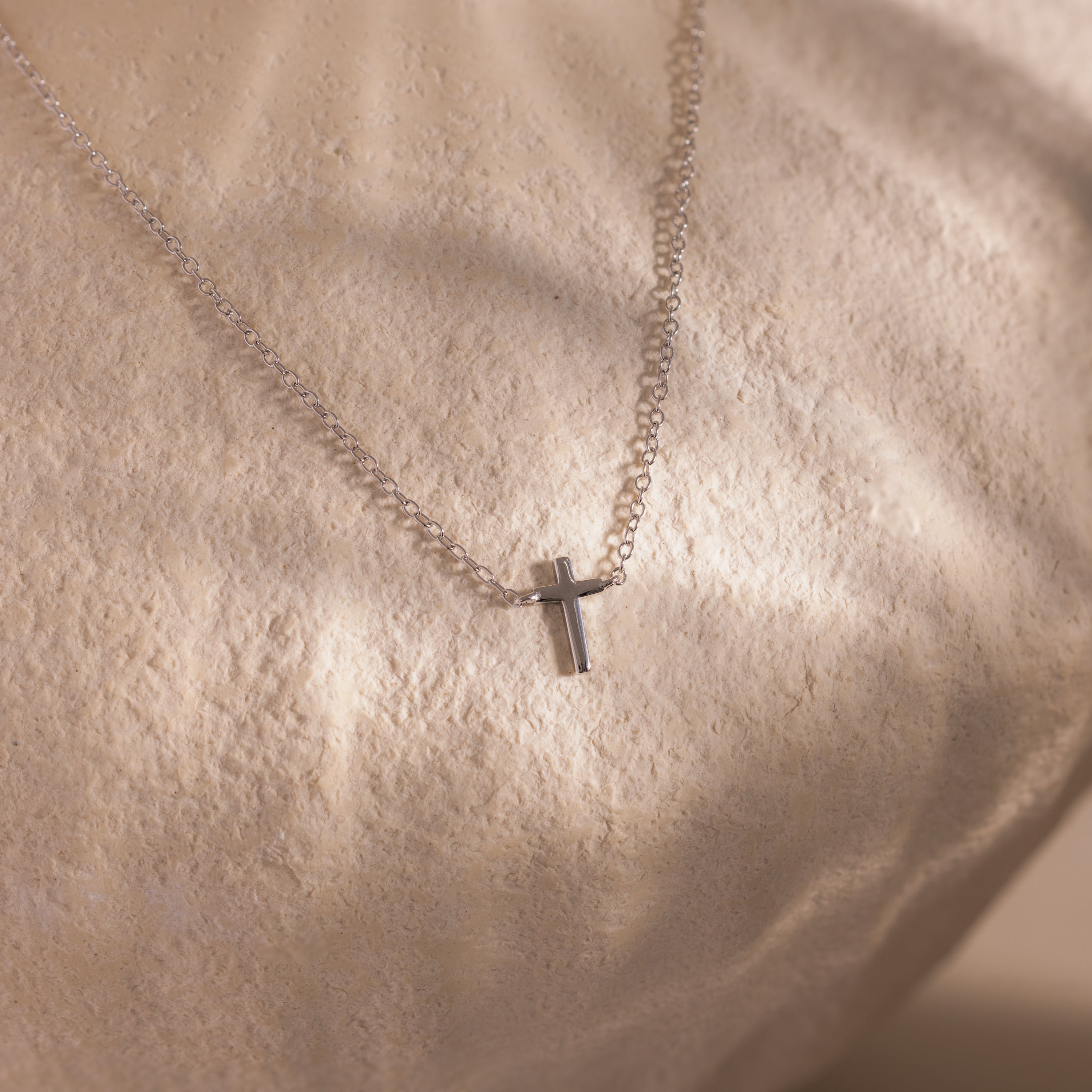 Dainty silver cross charm necklace displayed against a neutral stone surface.